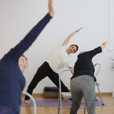 Tres personas en la postura del triángulo, adaptada con silla, durante una clase de Yoga Oncológico