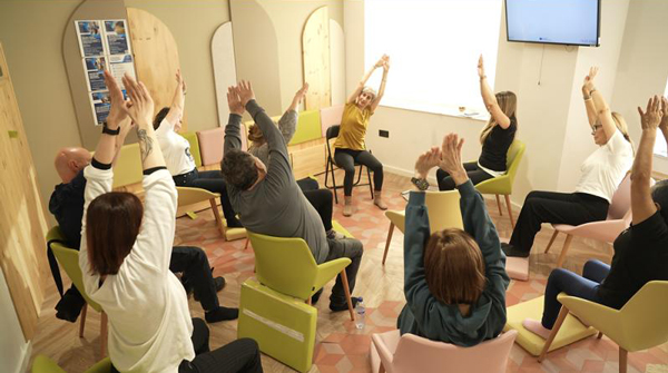 Grupo de yoga en sesión de yoga oncológico. Personas sentadas en sillas formando un círculo elevando los brazos.